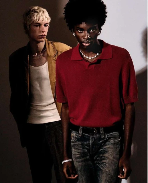 Two stylish men in casual attire pose indoors; one in a red polo shirt and jeans, the other in a white tank top and tan jacket.