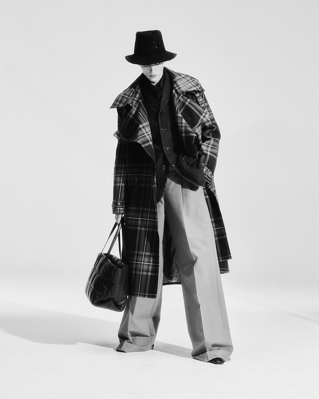 Person in a plaid coat, wide-leg pants, and hat, carrying a black bag, fashionably posing in a bright, neutral photo studio setting.