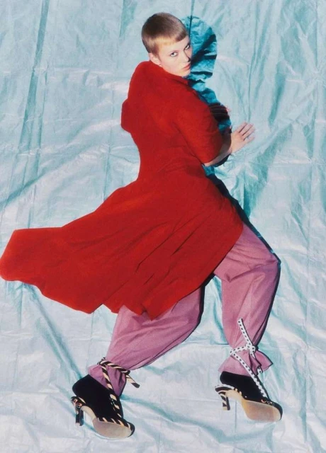Fashion photo of a person with short blonde hair, wearing a red dress, light pink pants, and high heels, lying on a light blue background.