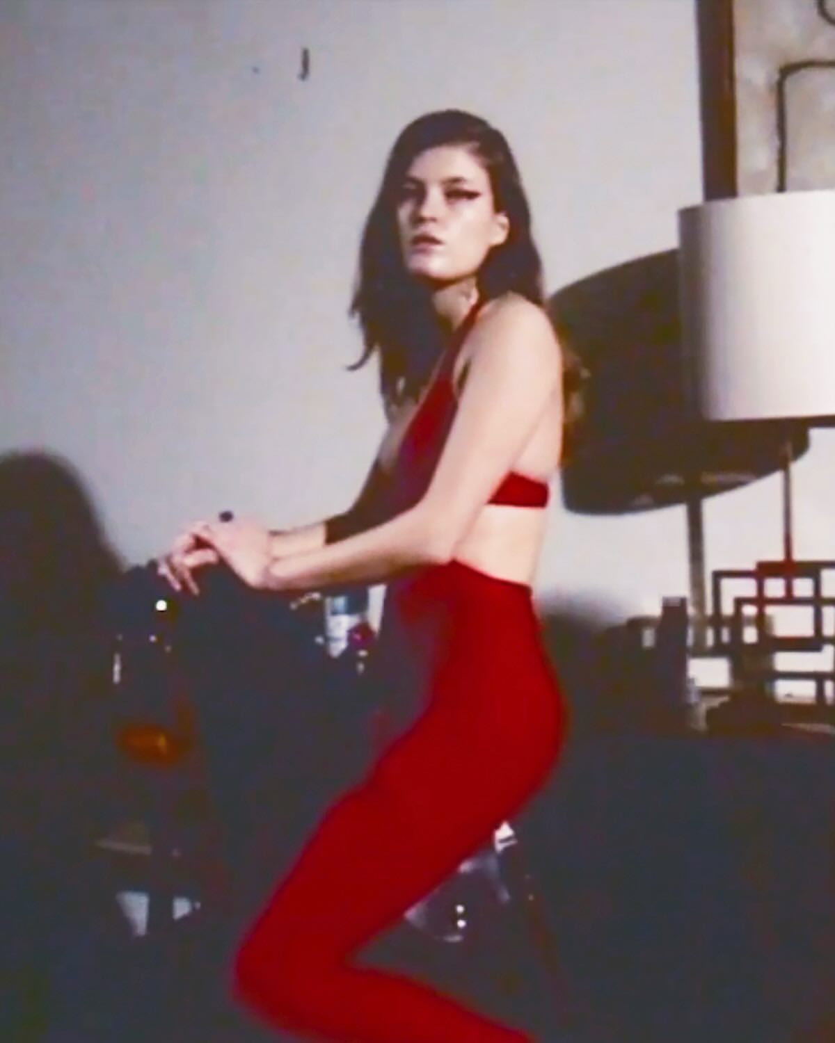 A woman in red athletic wear poses indoors near a black sofa, a standing lamp with a white shade, and a light grey wall.