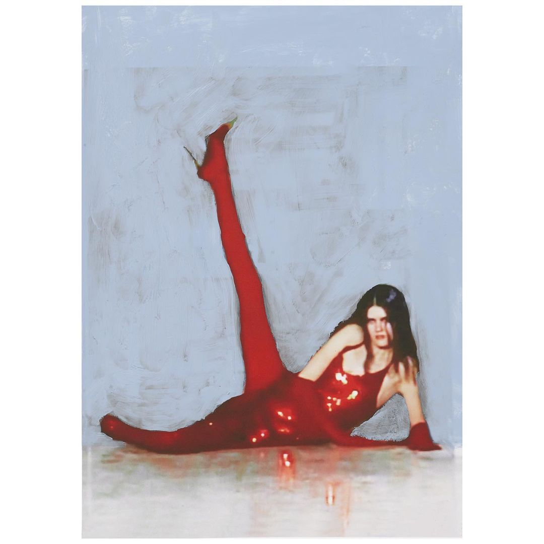 A woman poses while lying on the floor in a shiny red mini dress, red tights, and red high heels against a light blue background.