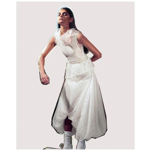 A woman poses wearing a sleeveless white dress with a tulle overlay, complemented by white boots, against a neutral backdrop in a studio setting.
