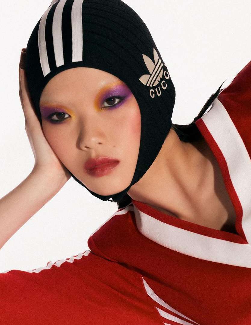 Close-up of an Asian woman wearing a black Adidas x Gucci knit hat, with colorful eye makeup and a red-and-white track suit.