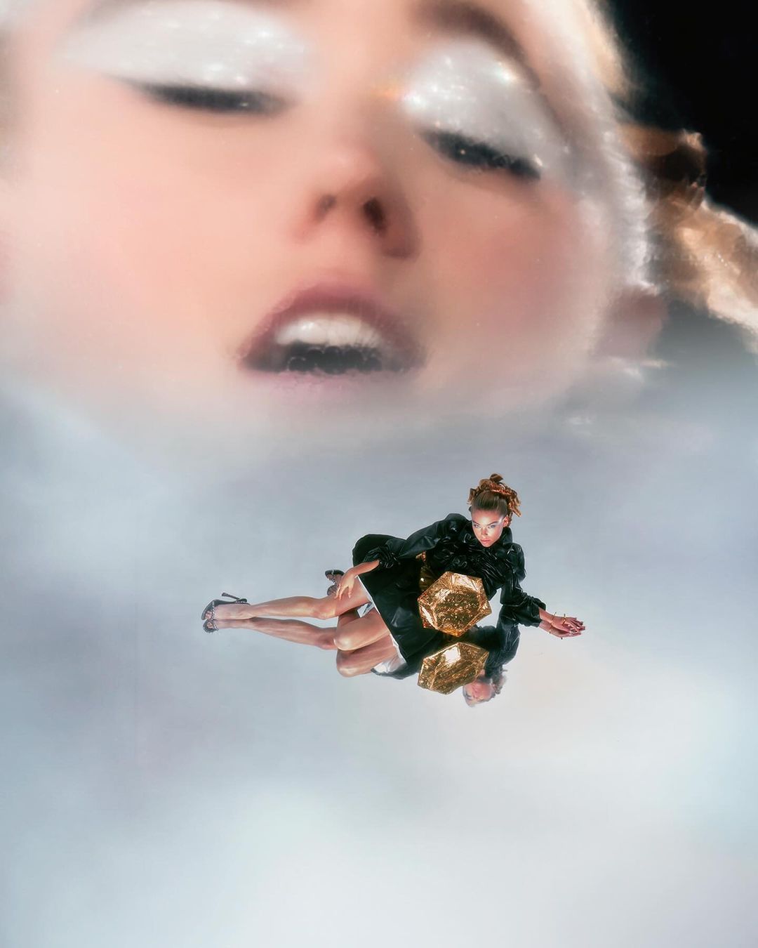 Fashion shot featuring a model lying on a reflective surface with a large face and white makeup visible in the background.