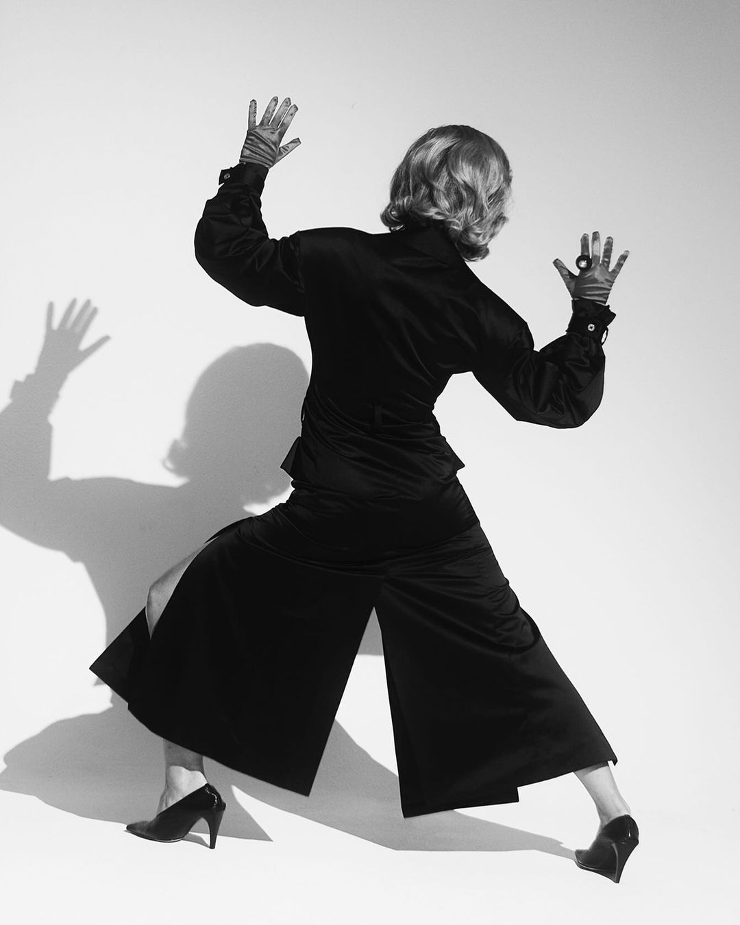 A person in black culottes and a matching top posing with arms up, the back view captured in a studio with a white backdrop.
