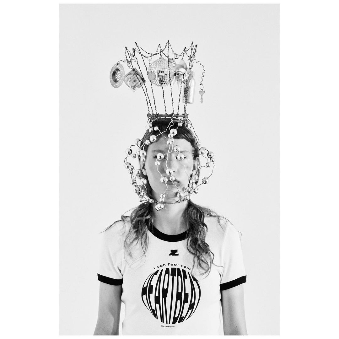 Black-and-white studio shot of a model wearing a pearl-covered face cage and a wire headdress holding miniature objects against a white backdrop.
