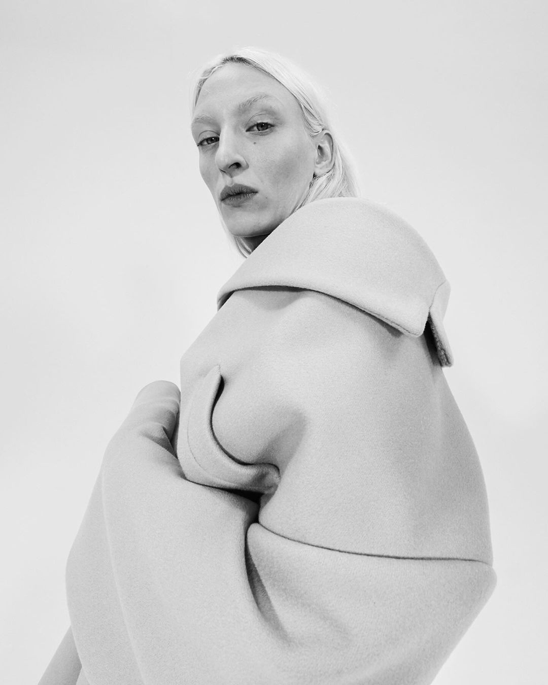 Monochrome fashion shot of a blond woman in an oversized cream coat posing in a photo studio with a bright white background, shoulder view.