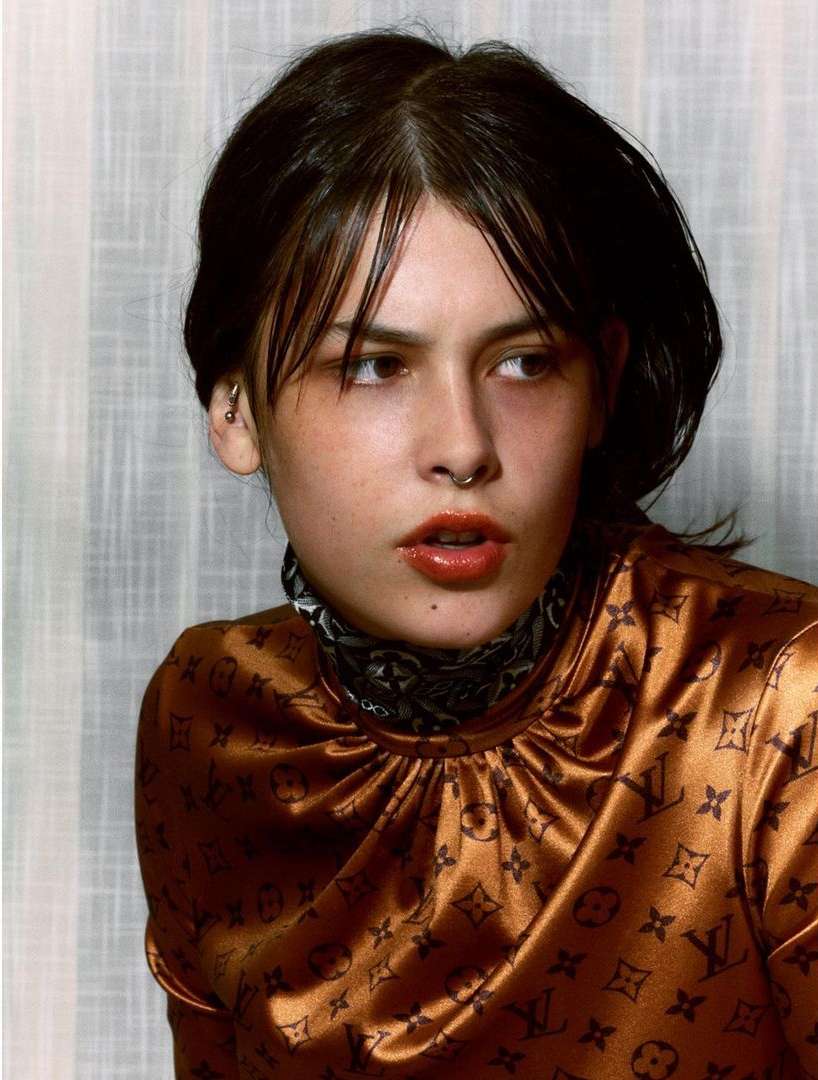 Close-up studio shot of a young adult woman with dark hair wearing a patterned top, looking off to the side against a striped backdrop.