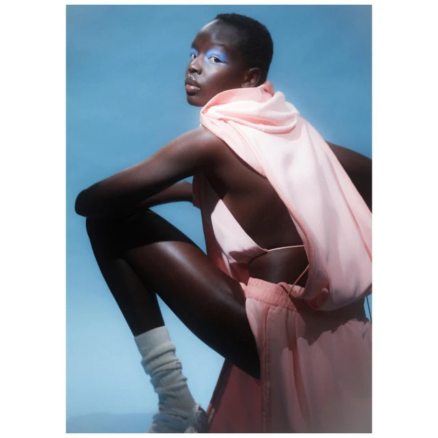 Portrait of a dark-skinned woman with blue eyeshadow in a pink outfit and white socks sitting against a bright blue background.