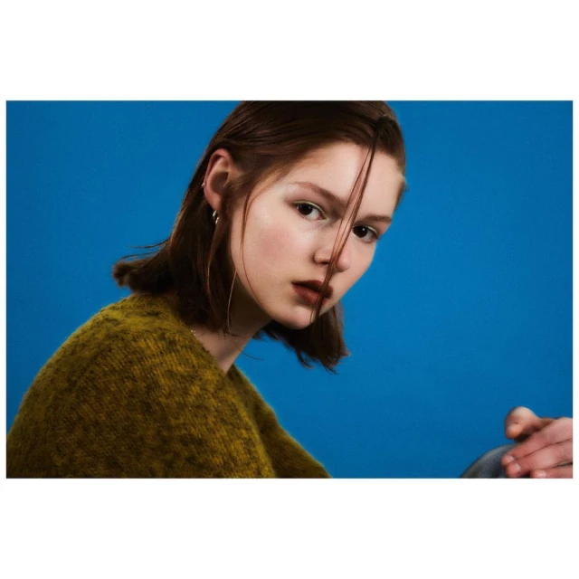 Portrait of a young woman with dark blonde hair and wearing an olive-green sweater against a solid blue background in a studio setting.