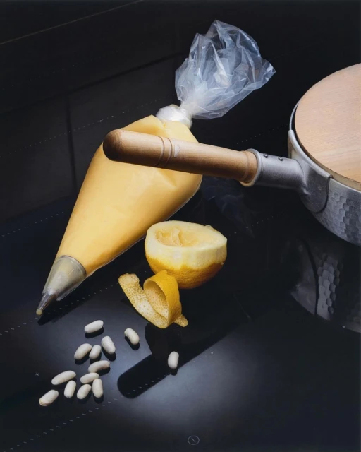 Pastry bag filled with yellow cream next to half a lemon and lemon peel, with scattered white candies on a glossy black surface.
