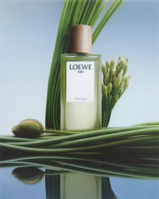 Loewe Aire Fantasia perfume bottle sits nestled among green foliage against a soft blue gradient background in a beauty product shot.
