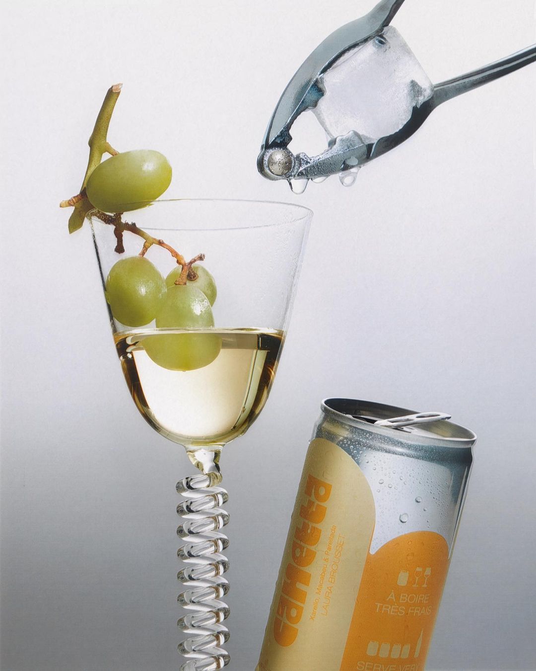 Still life shot of a wine glass filled with white wine and green grapes, alongside a silver can, with a pair of ice tongs.