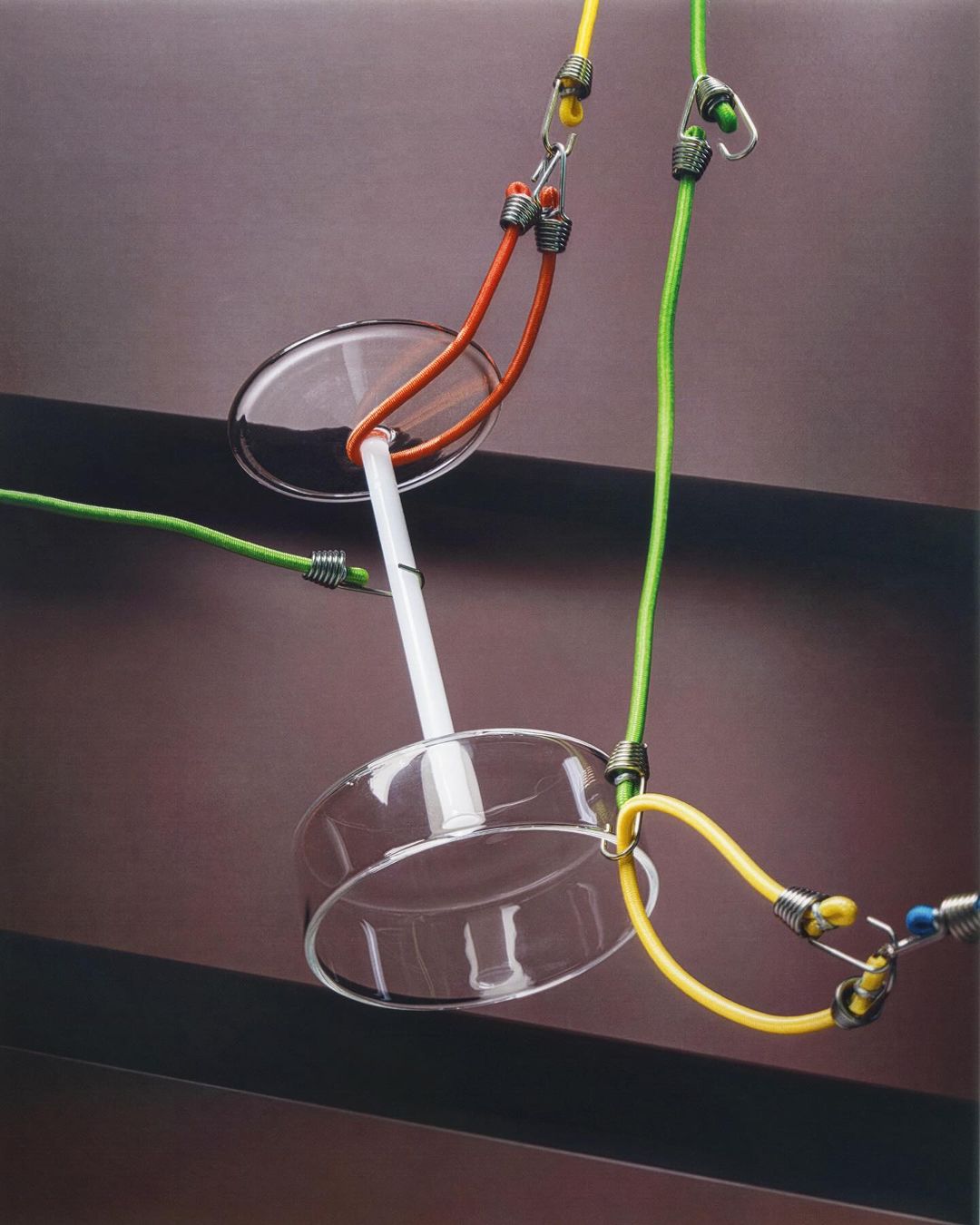 Still life of a drinking glass and colorful bungee cords against a brown background. The composition is clean and balanced.