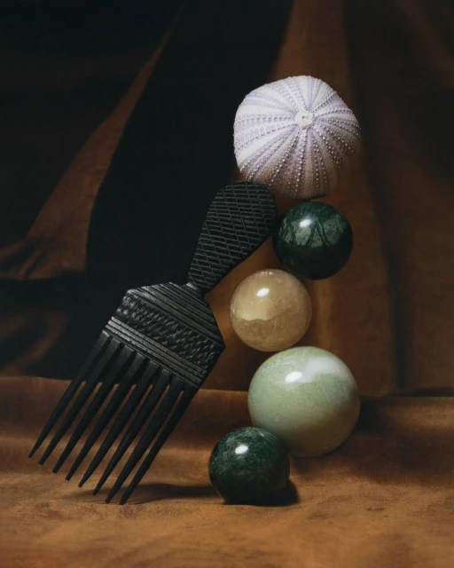 Still life photography featuring a black hair pick, a sea urchin, and marble-like spheres, artfully arranged on a brown fabric background.