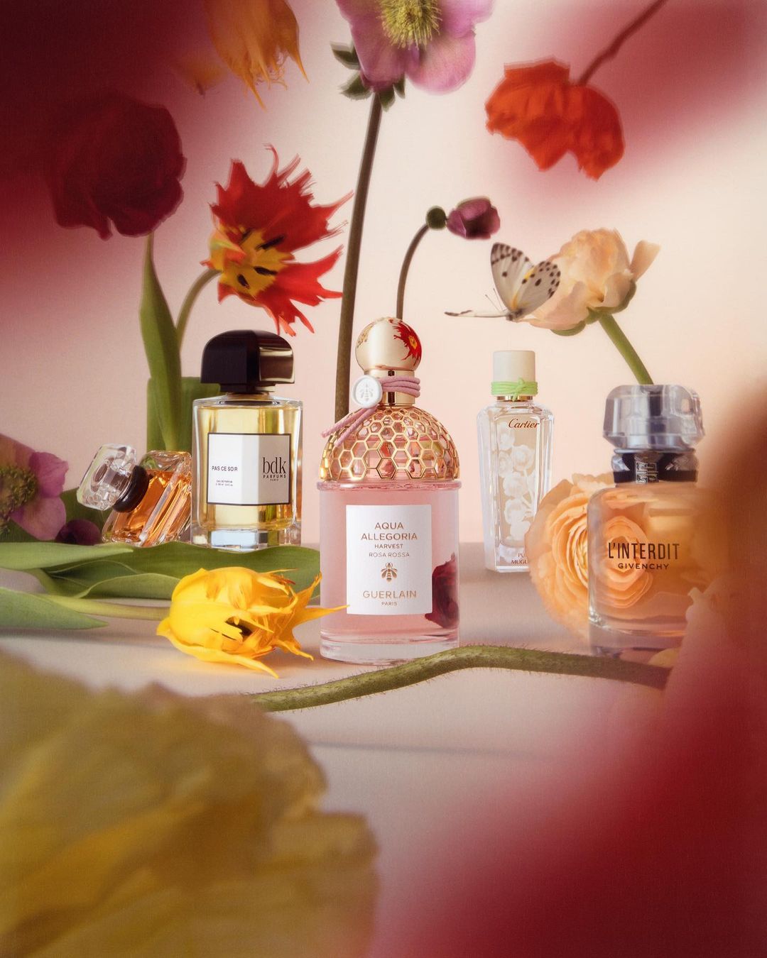 Five perfume bottles are displayed amongst colorful flowers with a butterfly on a blossom, with a soft pink background and diffused lighting.