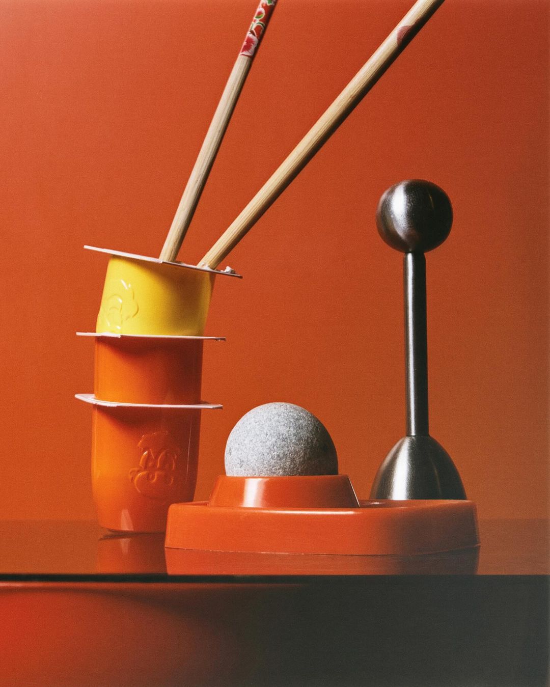 Still life photography of stacked containers and chopsticks next to a modern gray sphere on an orange reflective surface and backdrop.