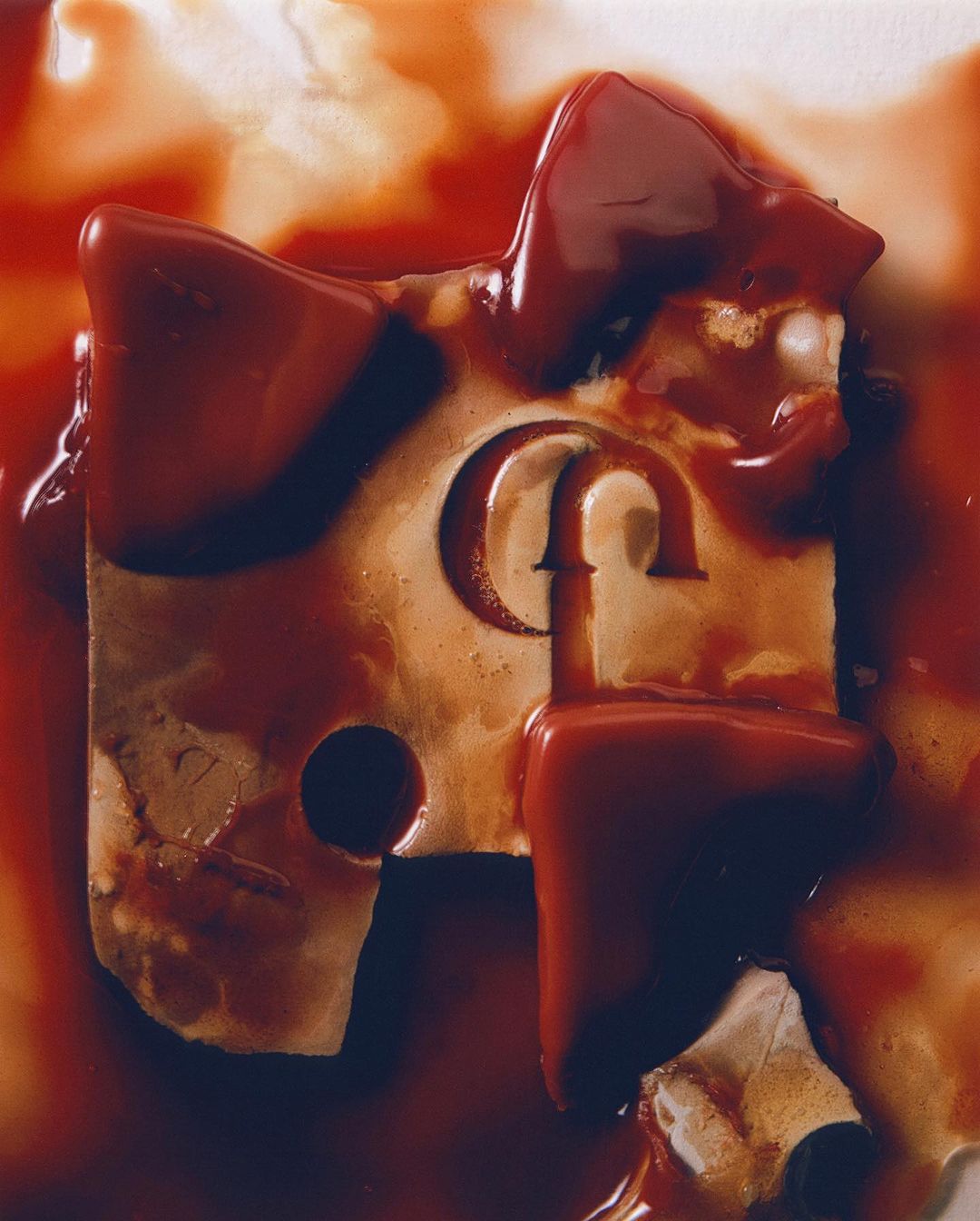 Abstract still life image featuring a piece of what appears to be processed cheese with a letter engraving and dripping red sauce.