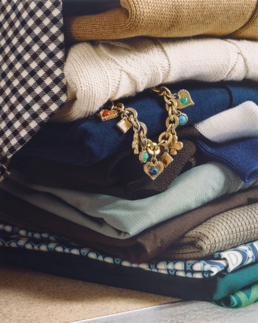 Neatly stacked sweaters and folded fabrics create a cozy scene. A gold charm bracelet drapes across a blue sweater.