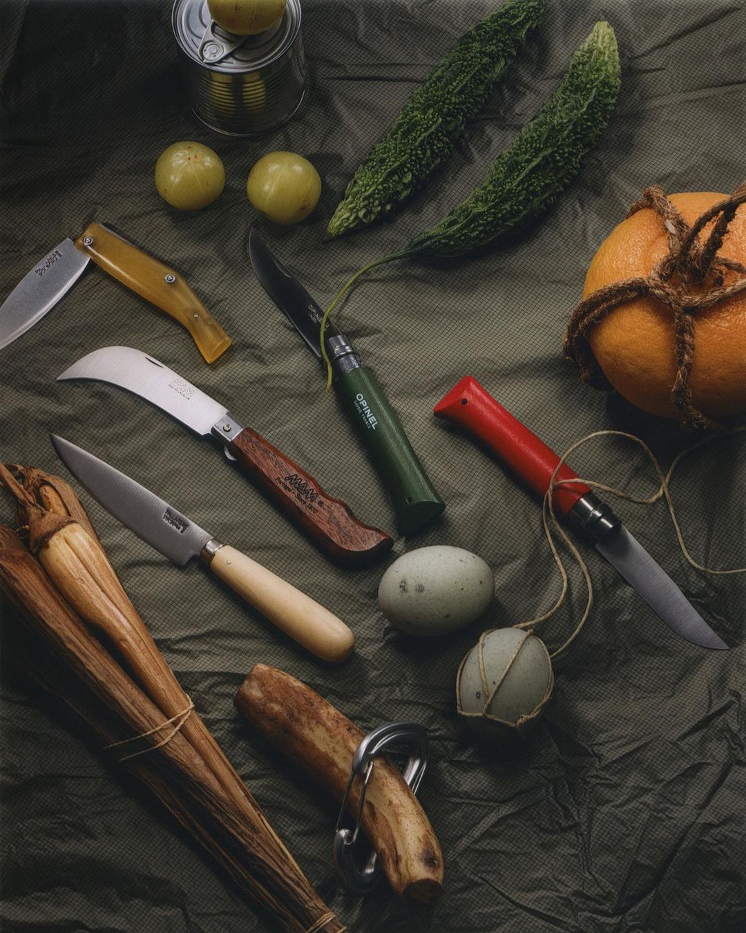 Still life featuring an assortment of knives, gourds, citrus fruits, wooden sticks, and eggs on a textured cloth, creating an artistic food display.