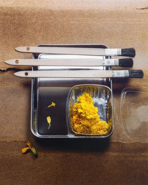 Overhead studio shot of brushes, a tray with liquid, and a container filled with yellow flowers against a textured brown backdrop.