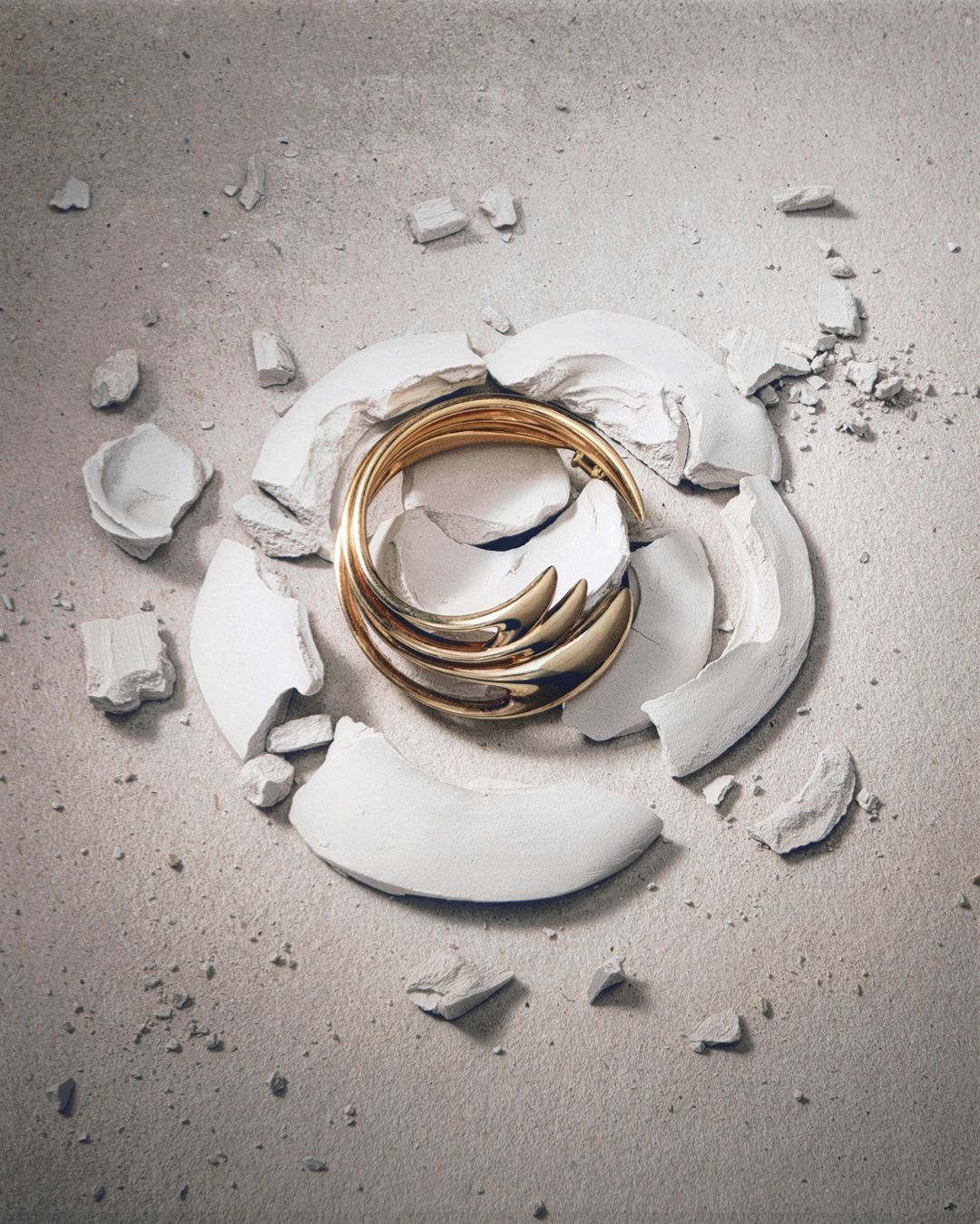 Abstract still life of gold bangle on a textured surface surrounded by broken white clay pieces, illuminated by a studio lighting setup.
