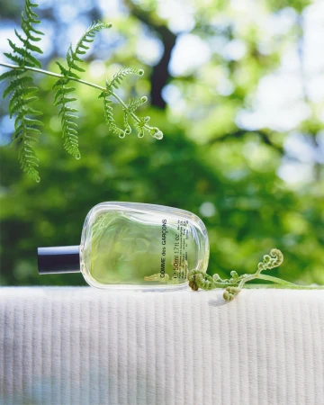 Bottle of Comme des Garçons perfume displayed on a white surface with fern fronds against a soft green bokeh background.