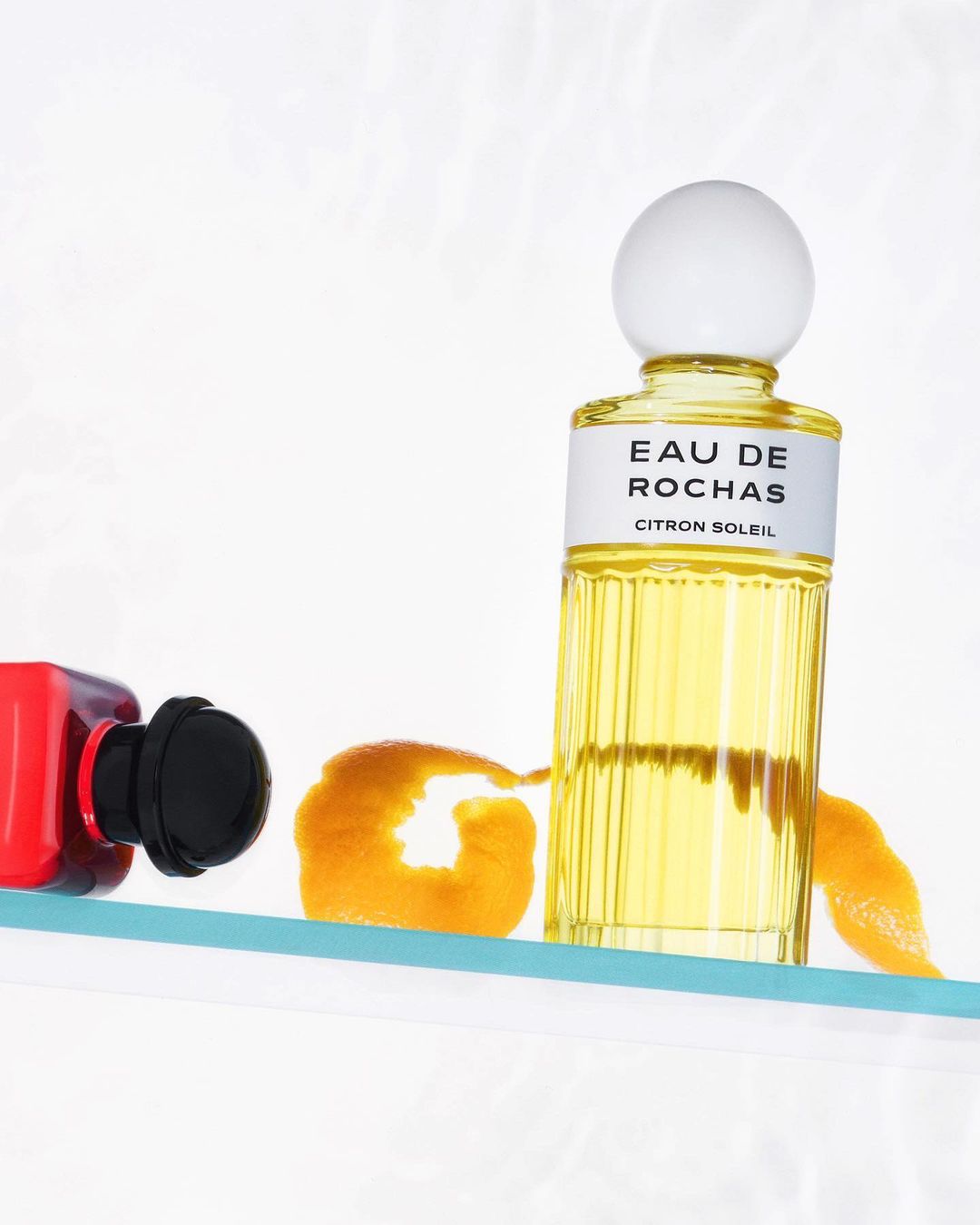 A bottle of Rochas Eau de Citron Soleil perfume stands next to an orange peel on a glass shelf against a white background in a studio shot.