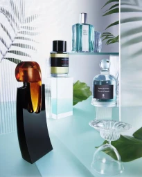Collection of glass perfume bottles displayed on tiered stands with green palm leaves and translucent panels in Paris by Adrien Sgandurra.