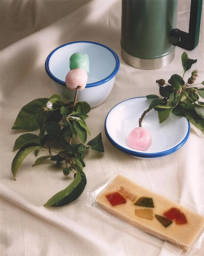 Still life photography featuring enamel bowls, green flask, leafy branches, mochi treats, and a bar of candy on fabric.