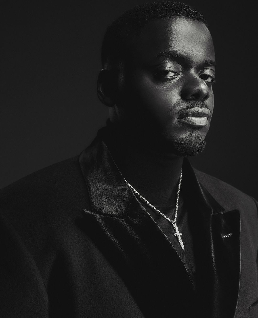 Portrait of Daniel Kaluuya wearing a dark blazer and necklace against a dark background by Adrienne Raquel