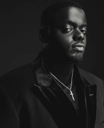 Portrait of Daniel Kaluuya wearing a dark blazer and necklace against a dark background by Adrienne Raquel