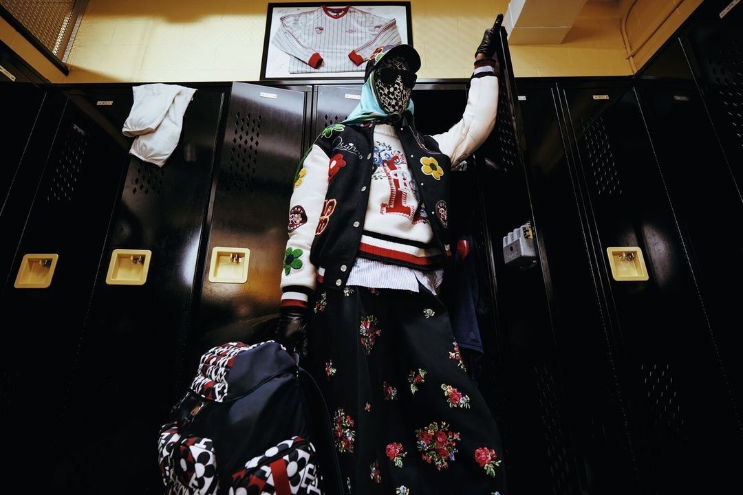 Person in patterned clothing and mask stands in front of black metal lockers in an indoor setting, holding a white sheet of paper.