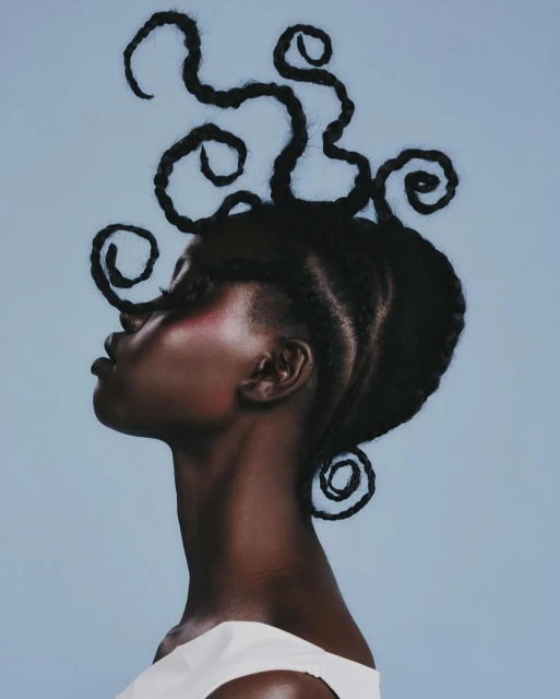 Profile portrait of a Black female model with intricate braided updo, creative coil extensions, and minimalist white top against light blue.
