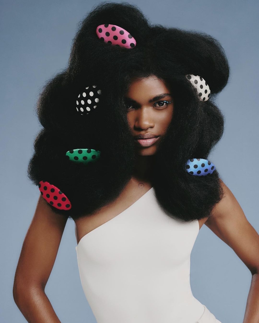Woman with afro hairstyle poses in a studio. The style features colorful polka dot hair clips. She is wearing a one-shoulder top.