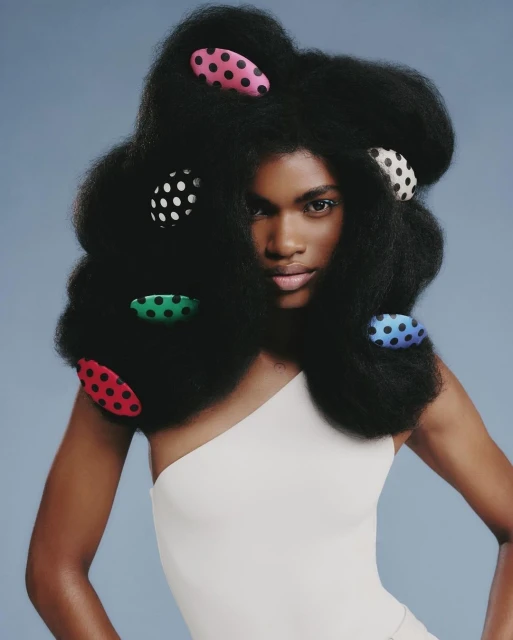 Woman with afro hairstyle poses in a studio. The style features colorful polka dot hair clips. She is wearing a one-shoulder top.