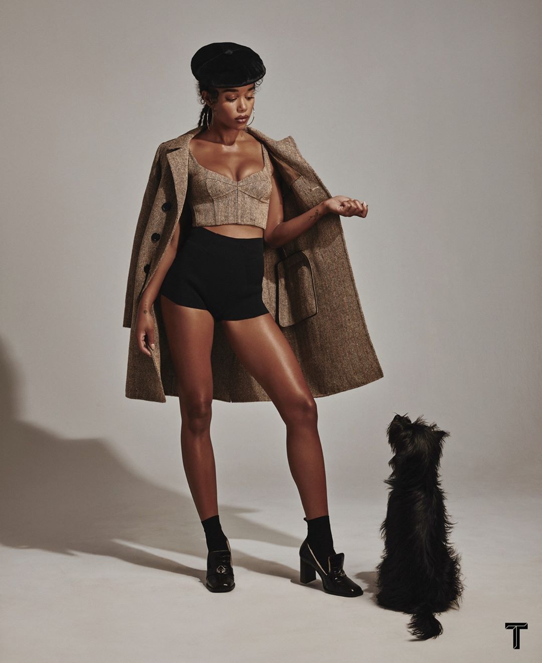 A woman poses indoors with a small dog, wearing a cropped top, shorts, heels, an overcoat and a beret in a studio setting.