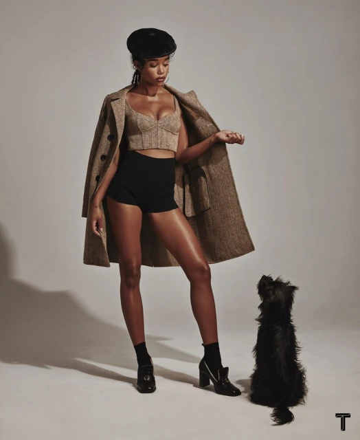 A woman poses indoors with a small dog, wearing a cropped top, shorts, heels, an overcoat and a beret in a studio setting.