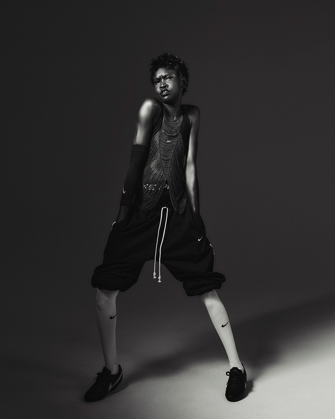 A person with short hair is posing in a studio, wearing a mesh top and shorts, with visible Nike logos on the leg and shoes, in black and white.