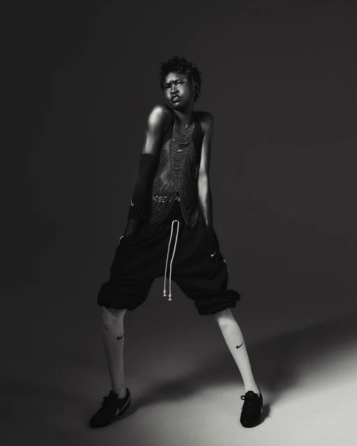 A person with short hair is posing in a studio, wearing a mesh top and shorts, with visible Nike logos on the leg and shoes, in black and white.