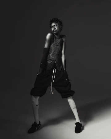 A person with short hair is posing in a studio, wearing a mesh top and shorts, with visible Nike logos on the leg and shoes, in black and white.