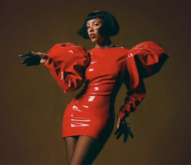 Glamorous singer in a glossy red dress and gloves poses indoors against a neutral background in a beauty and fashion shot.