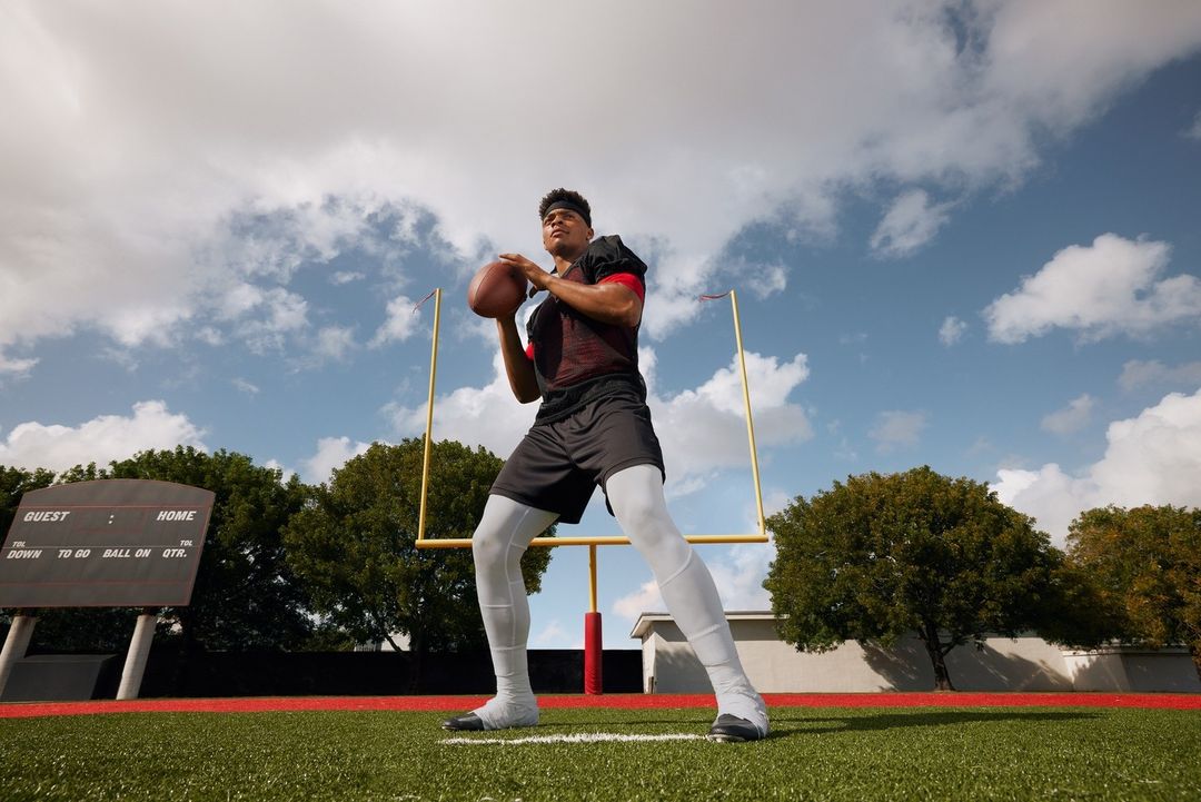 American football player throwing a football outdoors on a green field with a goal post and cloudy sky in the background.