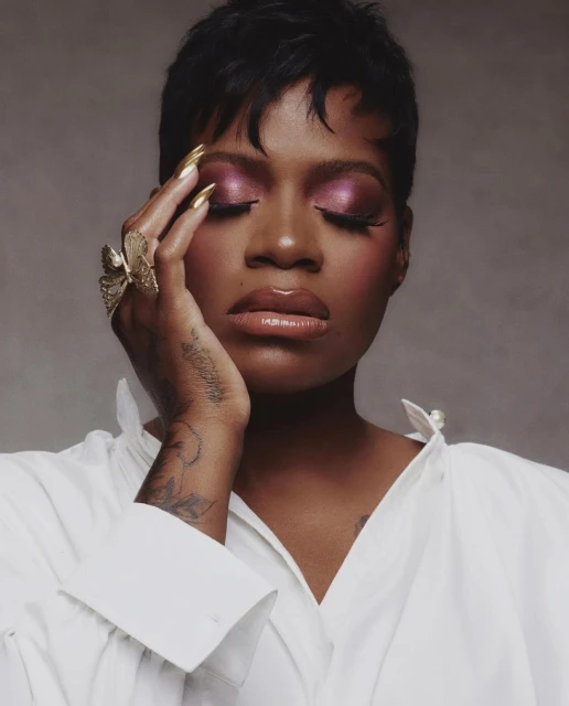 Close-up portrait of a woman with short dark hair, gold nails, and pink eyeshadow, posing with her eyes closed, wearing a white shirt.