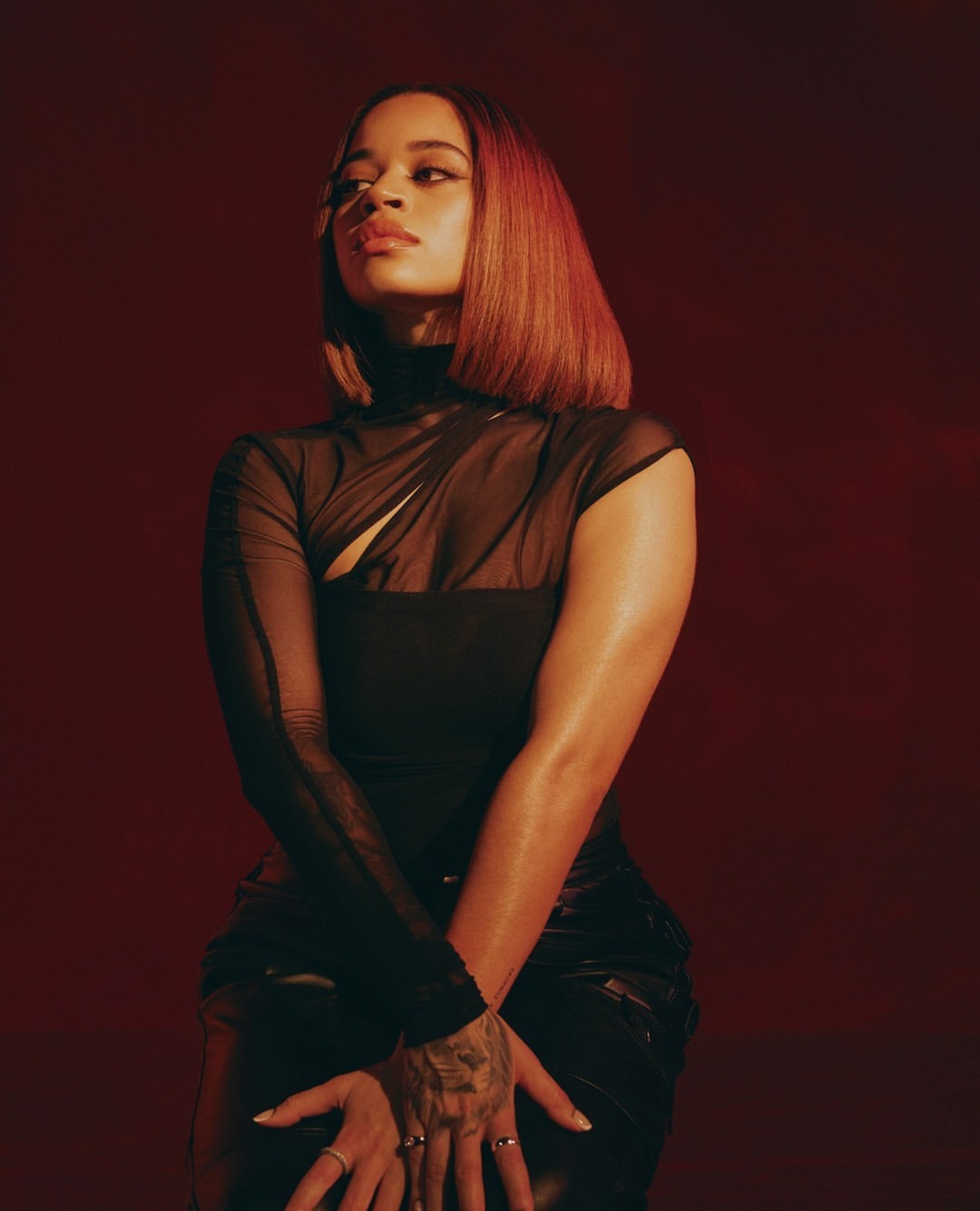 Portrait of a woman with auburn bob wearing a black, sheer asymmetrical top in front of a red background in a studio setting.