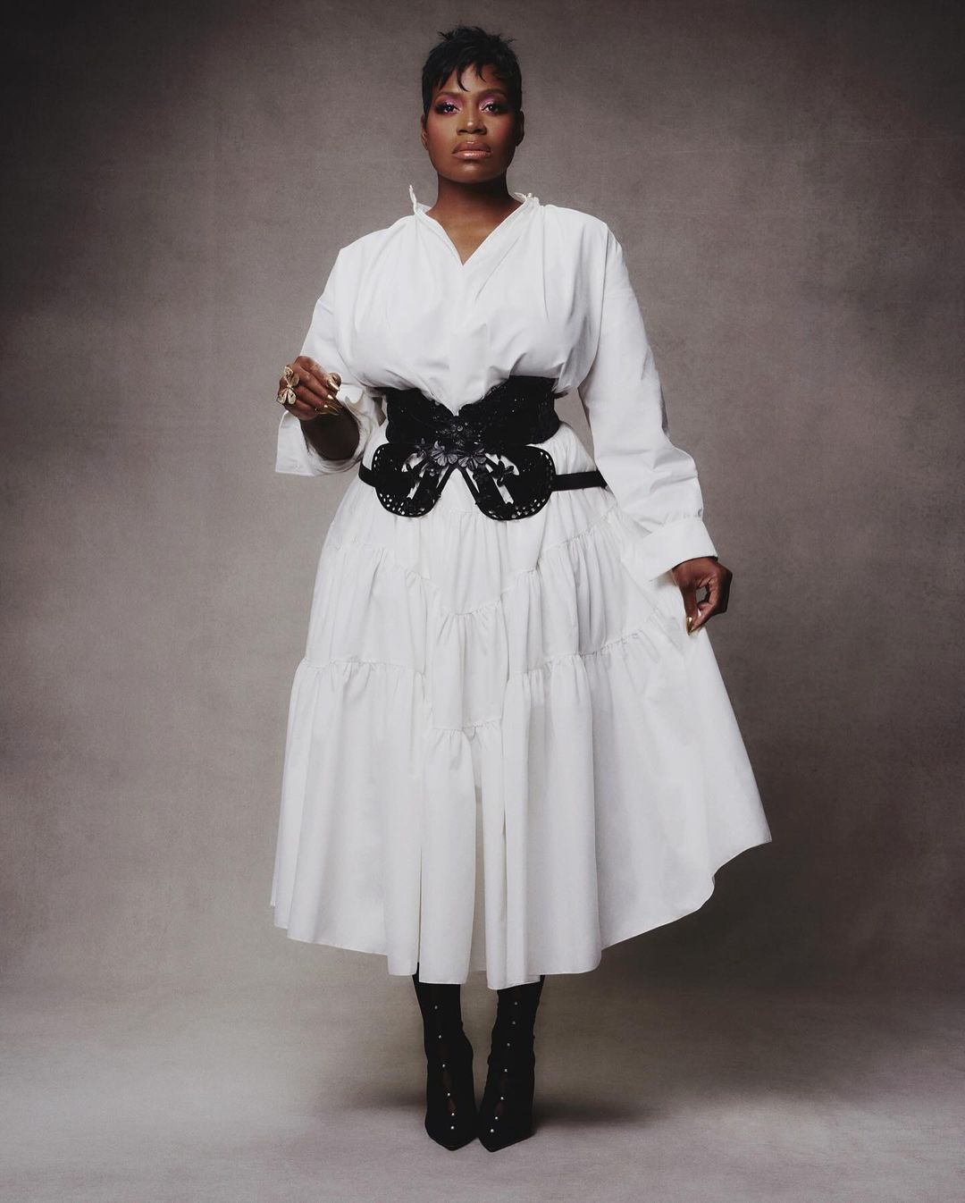 Full-length portrait of an African American woman in a white dress with a black corset, posing against a textured grey background.