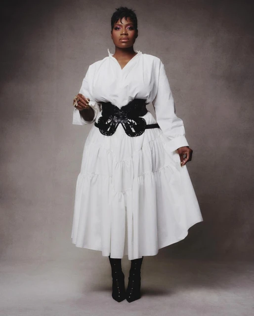 Full-length portrait of an African American woman in a white dress with a black corset, posing against a textured grey background.