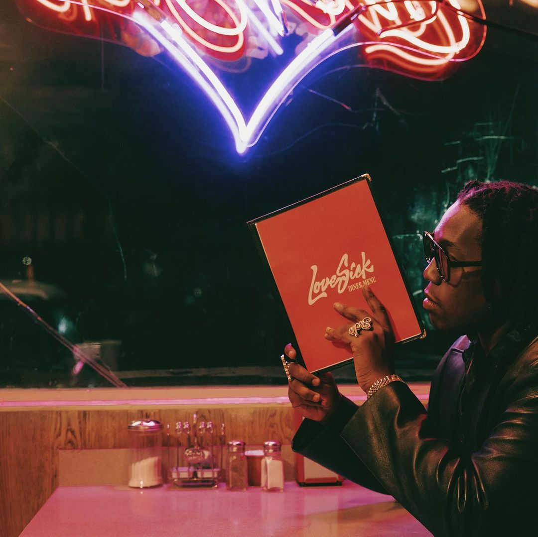 Adult male with Love Sick menu in retro diner setting, illuminated by neon heart. He wears glasses, leather jacket, gold jewelry.