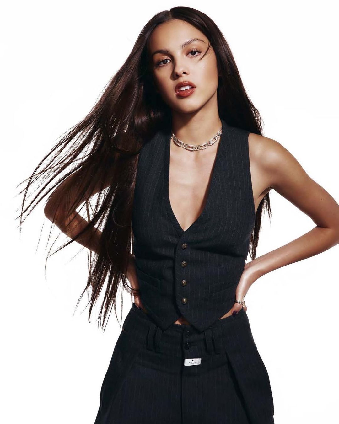 Portrait of a young woman with long brown hair, wearing a pinstripe vest and pants, posing against a white background in a studio shot.