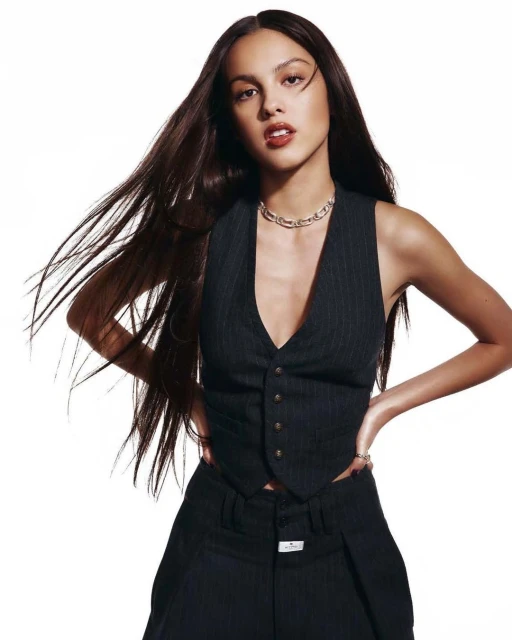 Portrait of a young woman with long brown hair, wearing a pinstripe vest and pants, posing against a white background in a studio shot.